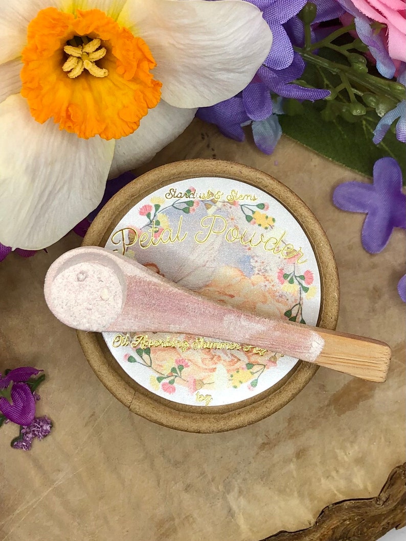 May include: A small, round, brown container with a white label that says "Stardust & Stems Petal Powder Oh, Apothecary Shimmer 0.5 oz". The container has a pink and white spoon with a wooden handle.