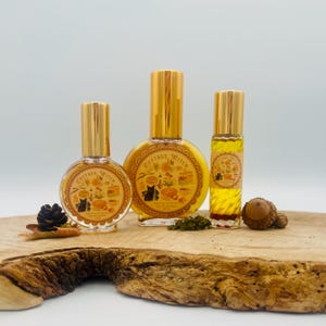 May include: Three glass perfume bottles with gold caps and round or cylindrical shapes. Each bottle features a circular label with the text Cottage Witch and illustrations of a black cat, food, and plants. The liquid inside is a golden yellow color.