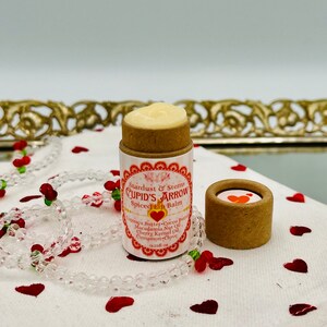Op de afbeelding: Een klein, bruin kartonnen buisje met een wit label waarop staat "Stardust & Stems CUPID'S ARROW Spiced Lip Balm". Het buisje is gevuld met een witte lippenbalsem en staat op een wit oppervlak met rode hartjes eromheen.