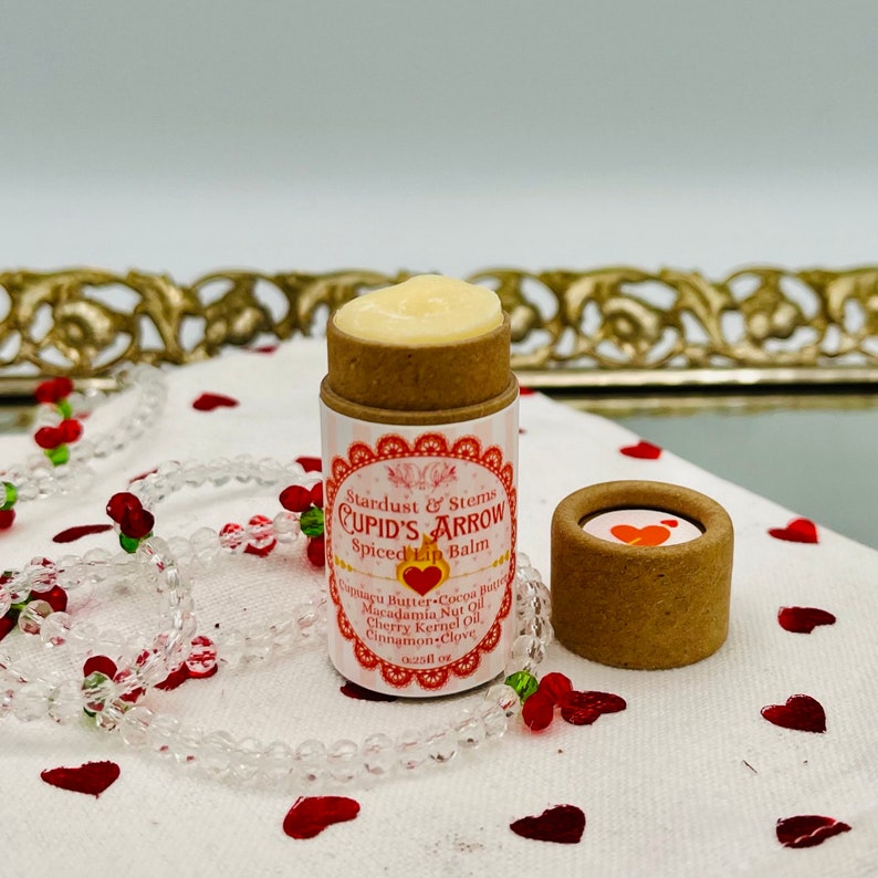 Op de afbeelding: Een klein, bruin kartonnen buisje met een wit label waarop staat "Stardust & Stems CUPID'S ARROW Spiced Lip Balm". Het buisje is gevuld met een witte lippenbalsem en staat op een wit oppervlak met rode hartjes eromheen.