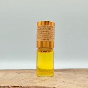 May include: A small clear glass vial filled with a golden yellow liquid, featuring a metallic gold cap and a beige paper label. The label text reads Stardust & Stems Cottage Witch, listing various scent notes and the word SAMPLE in bold.