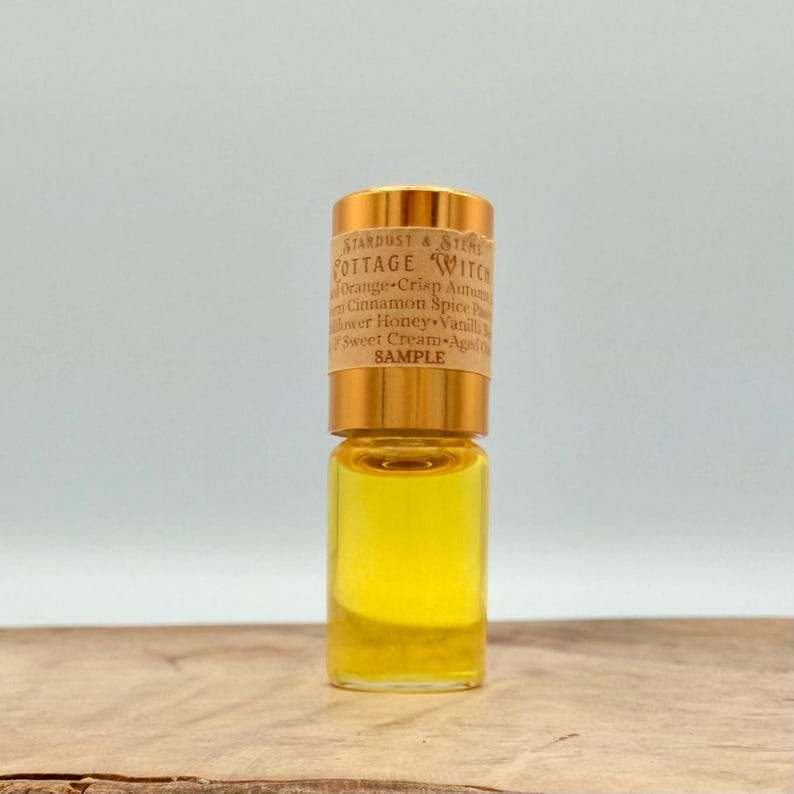May include: A small clear glass vial filled with a golden yellow liquid, featuring a metallic gold cap and a beige paper label. The label text reads Stardust & Stems Cottage Witch, listing various scent notes and the word SAMPLE in bold.