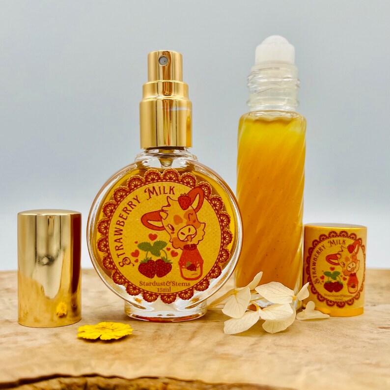 Puede incluir: Dos botellas de perfume con aroma a leche de fresa. La botella m&aacute;s grande es una botella de spray con una tapa dorada y una etiqueta redonda con una jirafa de dibujos animados y el texto "Strawberry Milk". La botella m&aacute;s peque&ntilde;a es una botella de roll-on con una tapa dorada y una etiqueta similar. Ambas botellas est&aacute;n sobre una superficie de madera con flores.