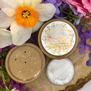 May include: A round, brown cardboard container with a white label that reads "Starlight Blooms Petal Powder Rose Petal & Minerals" with a vintage illustration of a woman in a white dress and flowers. The container has a matching lid with holes for dispensing powder. A white, fluffy puff is also included.