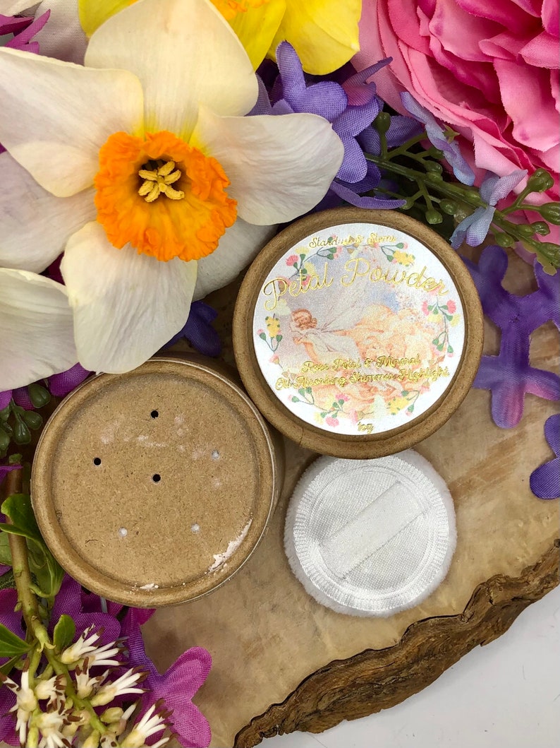 May include: A round, brown cardboard container with a white label that reads "Starlight Blooms Petal Powder Rose Petal & Minerals" with a vintage illustration of a woman in a white dress and flowers. The container has a matching lid with holes for dispensing powder. A white, fluffy puff is also included.