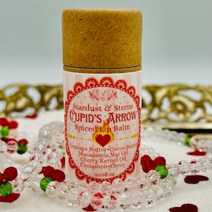Op de afbeelding: Een bruine kartonnen buis met een wit etiket waarop "Stardust & Stems CUPID'S ARROW Spiced Lip Balm" staat en de ingredi&euml;nten worden vermeld: "Cupuacu Butter, Cocoa Butter, Macadamia Nut Oil, Cherry Kernel Oil, Cinnamon Clove". Het etiket vermeldt ook "0.25fl oz".