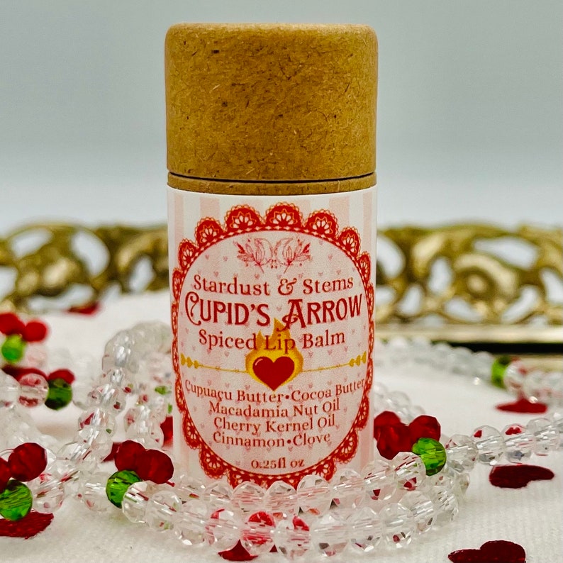 Op de afbeelding: Een bruine kartonnen buis met een wit etiket waarop "Stardust & Stems CUPID'S ARROW Spiced Lip Balm" staat en de ingredi&euml;nten worden vermeld: "Cupuacu Butter, Cocoa Butter, Macadamia Nut Oil, Cherry Kernel Oil, Cinnamon Clove". Het etiket vermeldt ook "0.25fl oz".