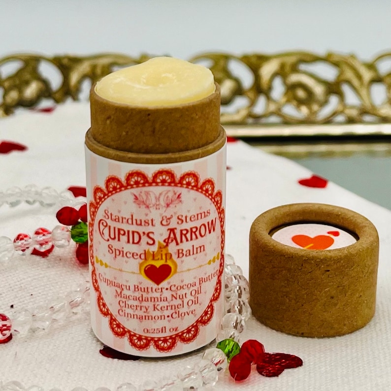 Op de afbeelding: Een kleine, bruine tube lippenbalsem met een wit label waarop staat "Stardust & Stems CUPID'S ARROW Spiced Lip Balm". Het label vermeldt ook de ingredi&euml;nten: "Cupuacu Butter, Cocoa Butter, Macadamia Nut Oil, Cherry Kernel Oil, Cinnamon-Clove". De tube is 0,25 fl oz.