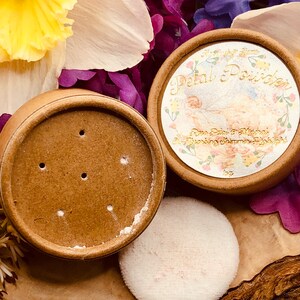 May include: A round, brown container with a white lid and a white puff. The container has the words "Petal Powder" printed on it in gold lettering. The container is surrounded by pink and purple flowers.