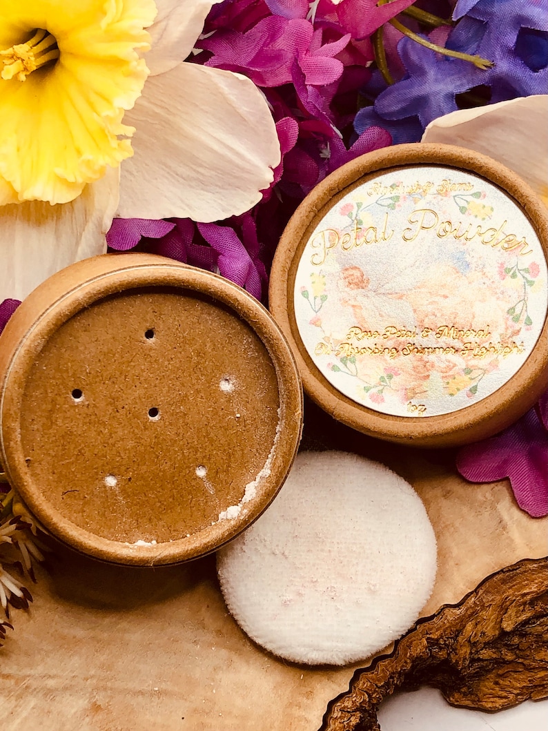 May include: A round, brown container with a white lid and a white puff. The container has the words "Petal Powder" printed on it in gold lettering. The container is surrounded by pink and purple flowers.