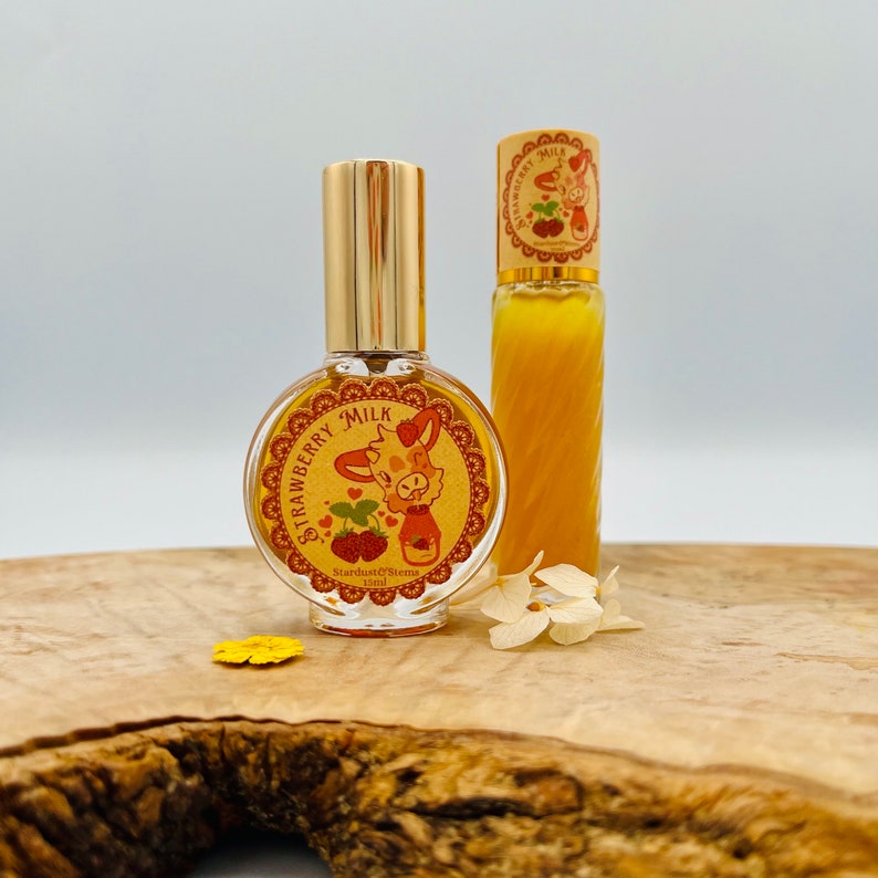 Puede incluir: Dos botellas de perfume de vidrio con tapas doradas y etiquetas. La botella de la izquierda tiene una etiqueta redonda con una jirafa de dibujos animados y el texto "Strawberry Milk" y "Stardust & Stems 10ml". La botella de la derecha tiene una etiqueta con el texto "Strawberry Milk". Ambas botellas est&aacute;n sobre una superficie de madera con flores blancas y amarillas.
