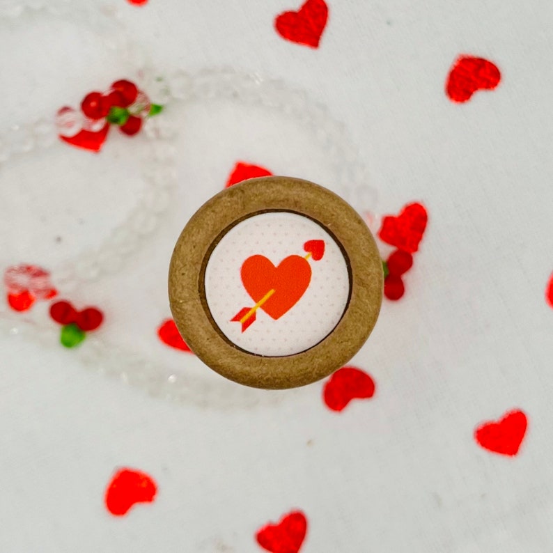 Op de afbeelding: Een houten knoop met een rood hart en pijl ontwerp. De knoop is omgeven door een witte achtergrond met rode harten.