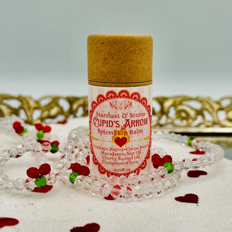 Op de afbeelding: Een bruine tube lippenbalsem met een roze en wit label waarop staat "Stardust & Stems CUPID'S ARROW Spiced Lip Balm". Het label vermeldt ook de ingredi&euml;nten: Cupuacu-boter, cacaoboter, macadamia-nootolie, kersenpitolie, kaneel-kruidnagel. De tube is 0,25 fl oz.
