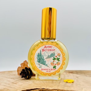 May include: A small glass bottle with a gold cap and a yellow label. The label has a picture of a snowy mountain scene with trees and the text "Alpine Buttercup" and "Stardust & Stems 1 fl oz".