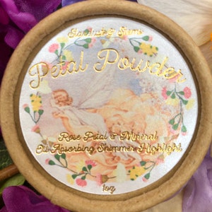 May include: A round, brown container with a white lid featuring a floral design and gold lettering. The lid reads "Stardust & Stems Petal Powder Rose Petal & Mineral Oil Absorbing Shimmer Highlight 10g".