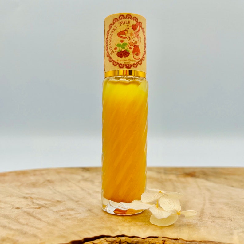 Puede incluir: Una botella de vidrio transparente con un l&iacute;quido amarillo en su interior. La botella tiene una tapa dorada con una fresa de dibujos animados y las palabras "Strawberry Milk" en ella. La botella est&aacute; sobre una superficie de madera con una flor blanca.