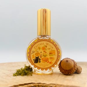 May include: A clear glass perfume bottle with a gold cap, labeled "Cottage Witch." The label features illustrations of a black cat, pie, and other whimsical elements. The bottle is displayed with moss and acorns on a wooden surface.