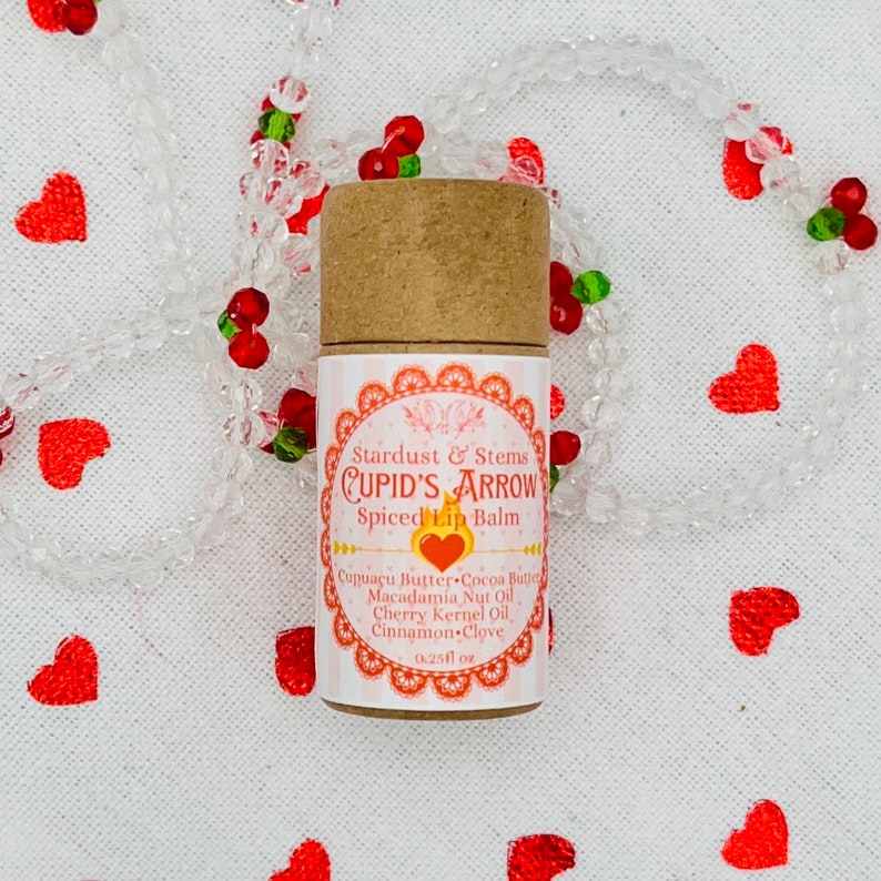Op de afbeelding: Een bruine tube lippenbalsem met een wit label waarop staat "Stardust & Stems CUPID'S ARROW Spiced Lip Balm" met een rood hart en de ingredi&euml;nten die eronder staan. De tube ligt op een witte achtergrond met rode harten en een heldere kralenketting.