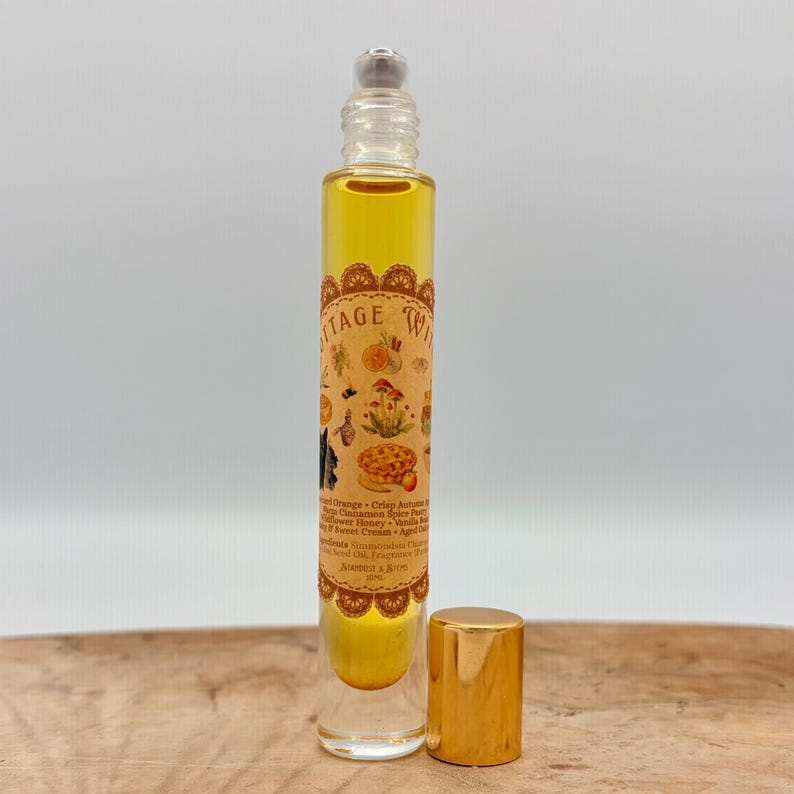 May include: A clear glass roll-on perfume bottle with a gold cap, containing golden liquid. The label features the text COTTAGE WITCH, illustrations of mushrooms, fruit, and a cat, and lists ingredients including Simmondsia Chinensis.