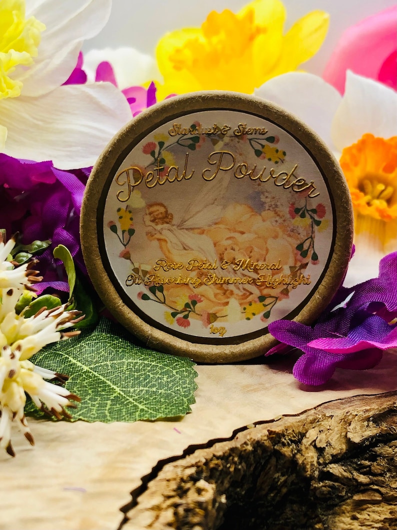 May include: A round, brown container with a gold lid and label. The label reads "Petal Powder Rose Petal & Mineral Oil Shimmering Highlight" and features a floral design with a fairy.
