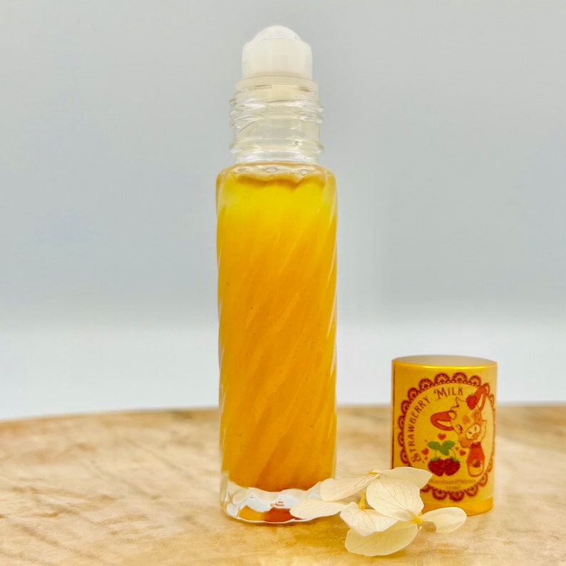 Puede incluir: Un frasco de vidrio transparente con un rodillo, que contiene un l&iacute;quido amarillo. El frasco tiene una bola de rodillo blanca en la parte superior y una tapa dorada con un dise&ntilde;o de leche de fresa y el texto "Strawberry Milk".