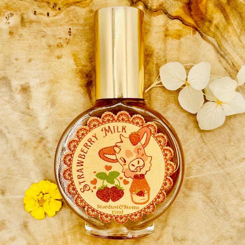 Puede incluir: Una botella redonda de vidrio con una tapa dorada y una etiqueta que dice "Strawberry Milk" con una ilustraci&oacute;n de dibujos animados de una vaca con una fresa en la cabeza. La etiqueta tambi&eacute;n dice "Stardust & Stems 15ml".