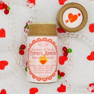 Op de afbeelding: Een bruine tube lippenbalsem met een wit label waarop staat "Stardust & Stems CUPID'S ARROW Spiced Lip Balm" en de ingredi&euml;nten worden vermeld: "Cupua&ccedil;u Butter Cocoa Butter Macadamia Nut Oil Cherry Kernel Oil Cinnamon Clove". Het label vermeldt ook "0.25 fl oz". De tube bevindt zich op een witte achtergrond met rode harten.