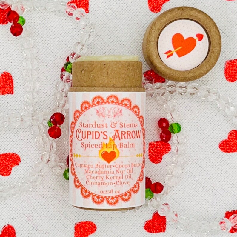 Op de afbeelding: Een bruine tube lippenbalsem met een wit label waarop staat "Stardust & Stems CUPID'S ARROW Spiced Lip Balm" en de ingredi&euml;nten worden vermeld: "Cupua&ccedil;u Butter Cocoa Butter Macadamia Nut Oil Cherry Kernel Oil Cinnamon Clove". Het label vermeldt ook "0.25 fl oz". De tube bevindt zich op een witte achtergrond met rode harten.