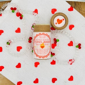 Op de afbeelding: Een bruine tube lippenbalsem met een roze en wit label waarop staat "Stardust & Stems CUPID'S ARROW Spiced Lip Balm". Het label vermeldt ook de ingredi&euml;nten: Cupuacu-boter, cacaoboter, macadamia-nootolie, kersenpitolie, kaneel-kruidnagel. De tube is omgeven door een draad van heldere kralen met rode harten en groene accenten.