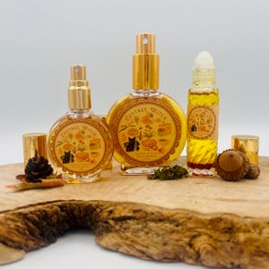May include: Three glass perfume bottles with gold caps and labels reading Cottage Witch. The labels feature illustrations of a black cat, mushrooms, and honey. The bottles contain a golden liquid and are arranged on a natural wood slice.