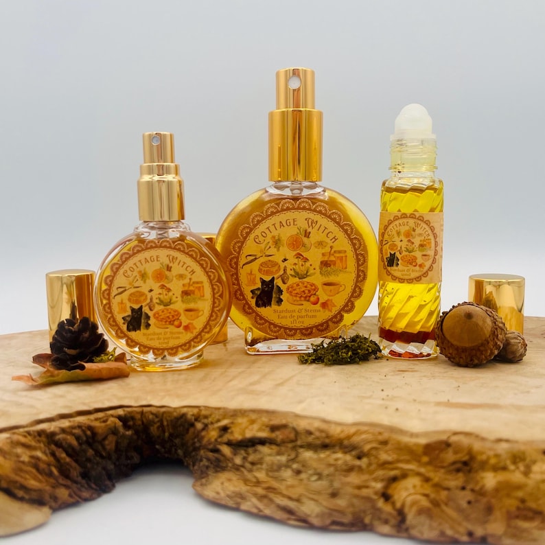 May include: Three glass perfume bottles with gold caps and labels reading Cottage Witch. The labels feature illustrations of a black cat, mushrooms, and honey. The bottles contain a golden liquid and are arranged on a natural wood slice.