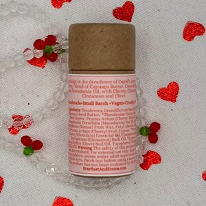 Op de afbeelding: Een bruine kartonnen buis met een roze etiket waarop de ingredi&euml;nten voor een veganistische lippenbalsem staan vermeld. Het etiket bevat de tekst "Cupid's Amorous Blend" en "Small Batch Vegan-Cruelty Free".