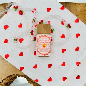 Op de afbeelding: Een bruine tube met een wit label waarop "Stardust & Stems CUPID'S ARROW Spiced Lip Balm" staat en de ingredi&euml;nten worden vermeld. De tube ligt op een witte stoffen achtergrond met rode harten.