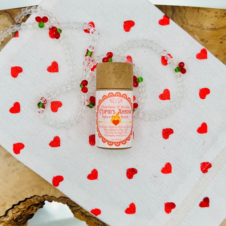 Op de afbeelding: Een bruine tube met een wit label waarop "Stardust & Stems CUPID'S ARROW Spiced Lip Balm" staat en de ingredi&euml;nten worden vermeld. De tube ligt op een witte stoffen achtergrond met rode harten.