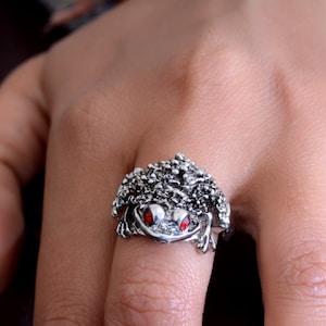 Puede incluir: Anillo plateado con forma de rana y ojos rojos. El anillo presenta un diseño detallado y texturizado en la cabeza y el cuerpo de la rana. La boca de la rana está ligeramente abierta.