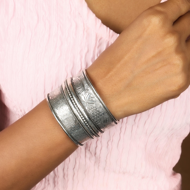 May include: A set of silver-toned metal cuff bracelets with intricate floral and geometric patterns. The bracelets are wide and worn on the wrist, showcasing detailed embossed designs. The jewelry is displayed against a soft pink background.