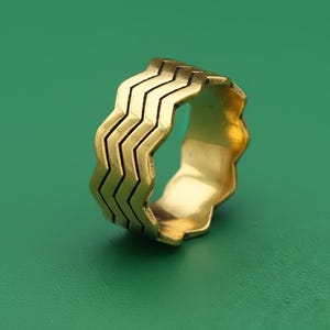 May include: A gold ring featuring a zigzag design around the band. The ring has a wavy edge and a polished finish. The ring is set against a green background.
