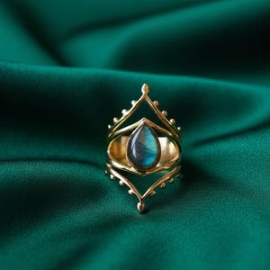 May include: A gold-colored ring with a teardrop-shaped, iridescent blue-green stone. The ring has an ornate, openwork design with a crown-like top and delicate details. The ring is set against a dark green, silky background.