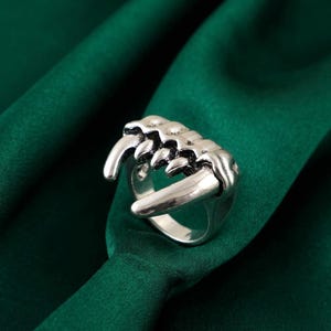 May include: A silver-colored ring featuring a claw design with multiple sharp points. The ring is set against a dark green background, highlighting its metallic sheen and intricate details. The claw-shaped design gives the ring a bold and edgy aesthetic.