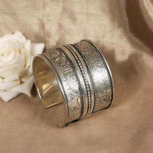 May include: Silver cuff bracelet with intricate floral and leaf patterns. The wide band features detailed engravings and a polished finish. The open design allows for easy wear. A decorative piece of jewelry.