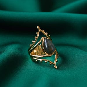 May include: A gold-colored ring with a teardrop-shaped, dark gray gemstone. The ring has an open, ornate design with decorative elements. The ring is set against a dark green background.
