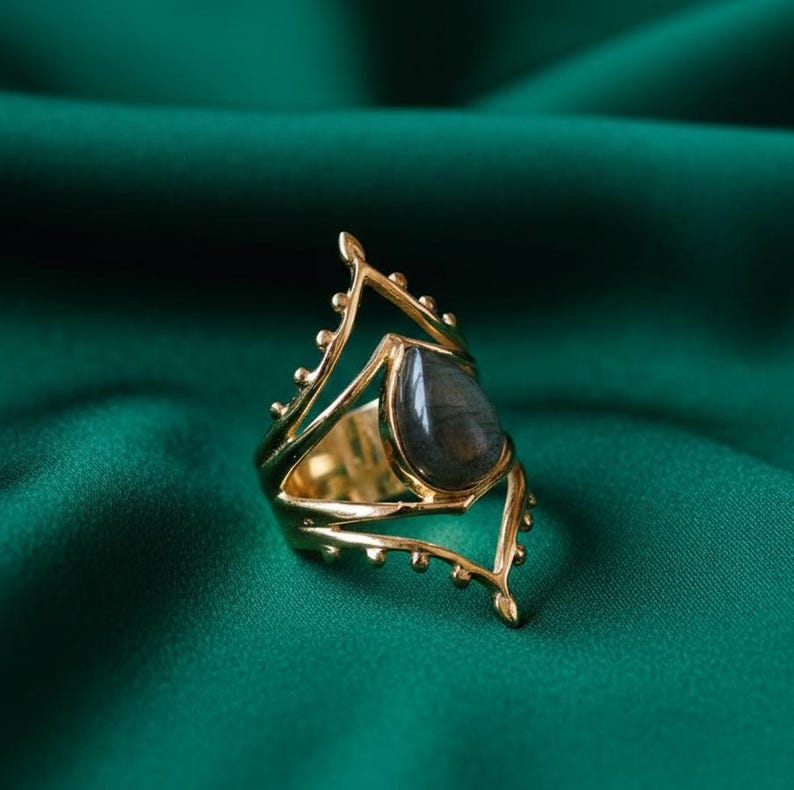 May include: A gold-colored ring with a teardrop-shaped, dark gray gemstone. The ring has an open, ornate design with decorative elements. The ring is set against a dark green background.