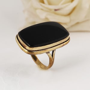 May include: A vintage ring featuring a large, rectangular black stone set in a gold-toned metal band. The ring has a classic design with a double-layered bezel around the stone. The band has a split shank design.