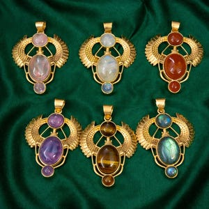 May include: Six gold-colored pendant necklaces with various gemstone inlays, including rose quartz, moonstone, carnelian, amethyst, tiger's eye, and labradorite. Each pendant features a stylized wing design. The background is a dark green fabric.