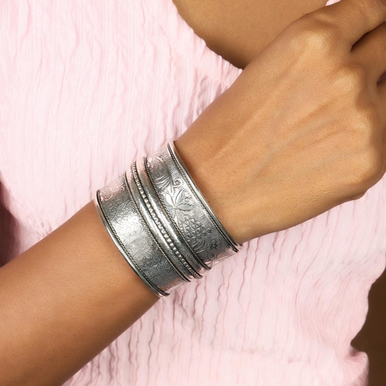 May include: A set of silver-toned metal cuff bracelets with intricate floral and geometric patterns. The bracelets are wide and stack together, creating a bold statement piece. The design includes detailed engravings and beaded accents.