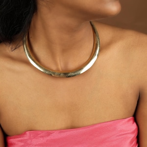 Puede incluir: Un collar dorado y curvo. El collar tiene un acabado liso y pulido y se lleva alrededor del cuello. El fondo es de color marrón liso. La persona lleva un top rosa sin tirantes.
