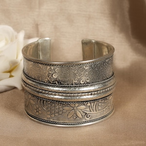 May include: Silver-toned cuff bracelet with intricate floral and paisley patterns. The wide band features detailed engravings and is accented with rope-like trim. The open design allows for easy wear. A decorative piece of jewelry.