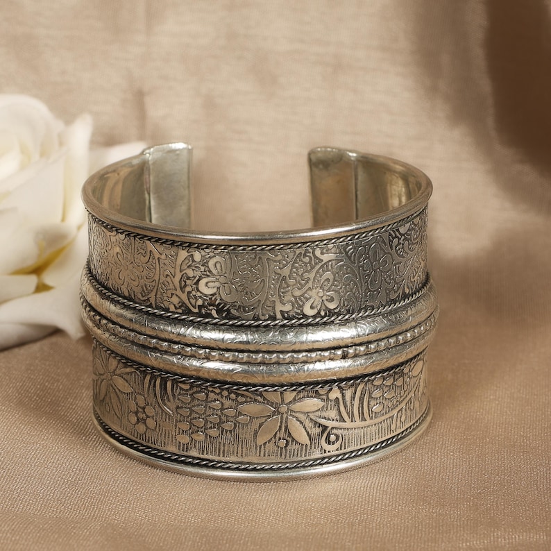 May include: Silver-toned cuff bracelet with intricate floral and paisley patterns. The wide band features detailed engravings and is accented with rope-like trim. The open design allows for easy wear. A decorative piece of jewelry.