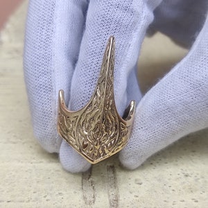 May include: A gold-colored, ornate pendant with a stylized, pointed design. The pendant features intricate, engraved patterns and a curved base with two small, upward-facing hooks. The overall shape is reminiscent of a stylized anchor or arrowhead.