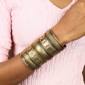 May include: Several gold-coloured bangles with detailed embossed patterns. The bangles are stacked on a wrist, displaying floral and geometric designs. The overall style is vintage and bohemian, with a warm, antique finish.